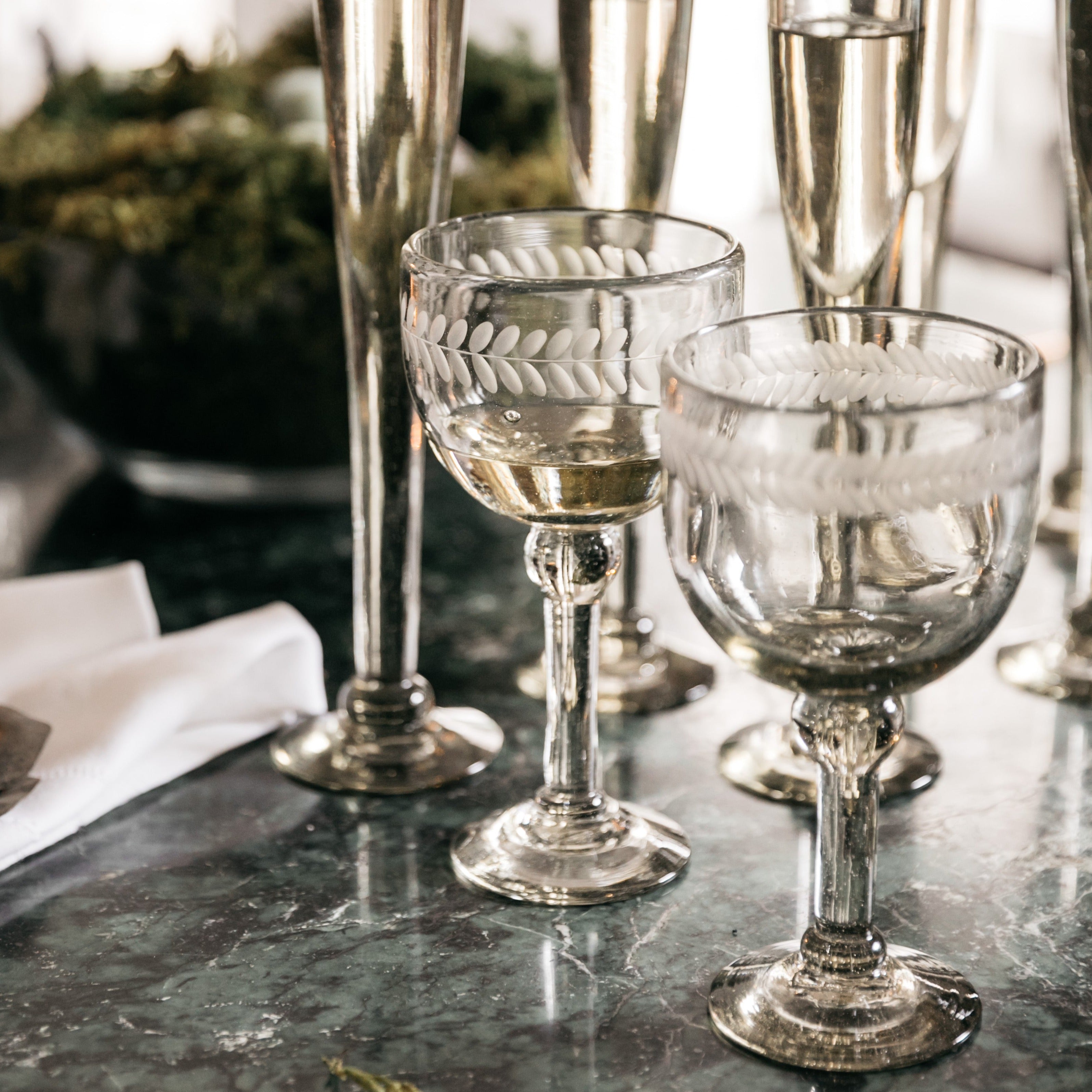Two clear mouth blown goblets with laurel engraving. In the background are several clear glass flutes.