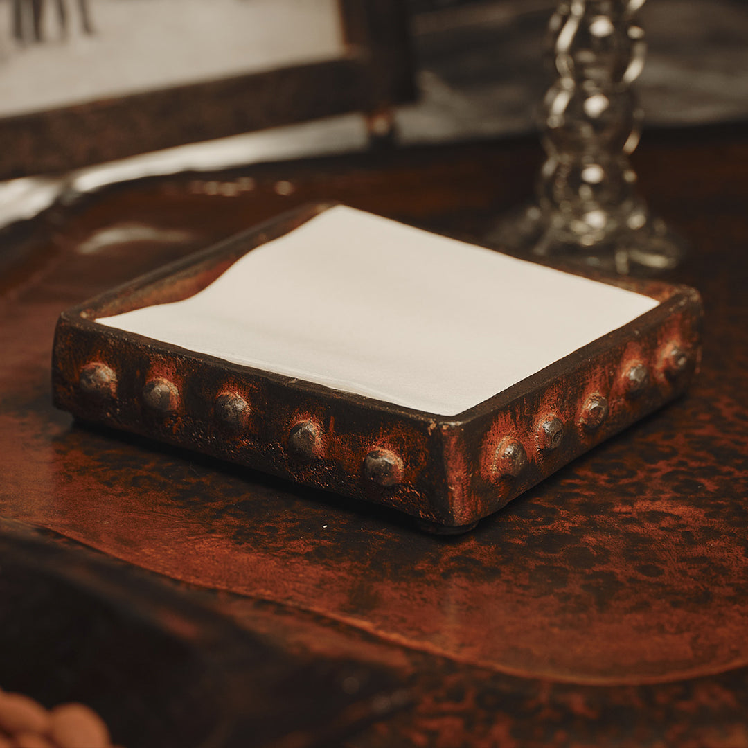 A wax cast iron square napkin holder with beaded detail trim. White napkins inside of it. It is resting on top of an iron surface.