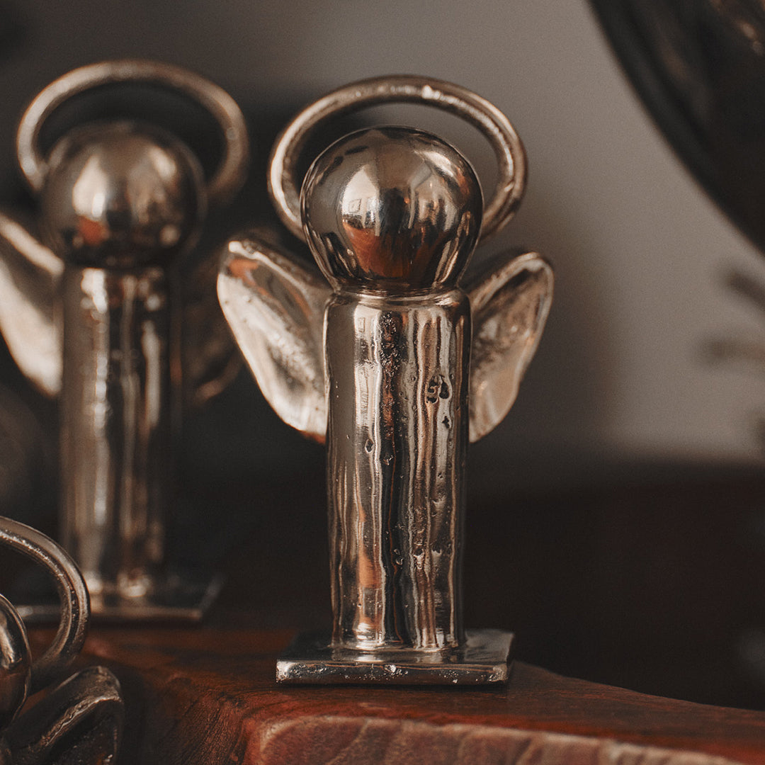 Two mini angels plated in nickel with halo on a square base on top of a wooden surface.