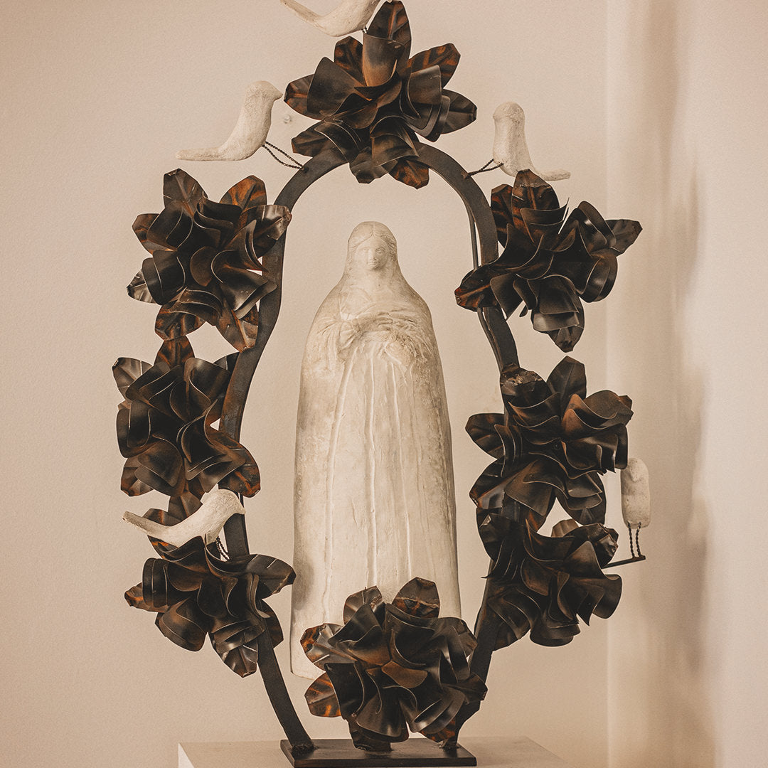 A hand carved stone statue representing the spirit of the virgin of Guadalupe surrounded by an aura and halo of iron flowers and stone birds. White wall background.