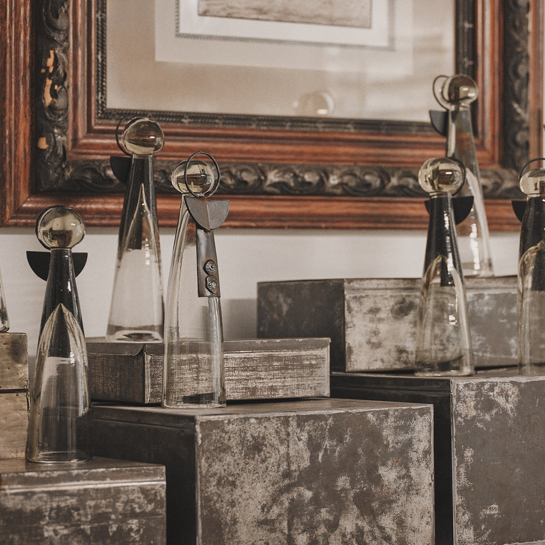 Several mouth blown glass angels with iron wings and halo standing next to each other on top of a grey and white surface. On the background is a brown frame.