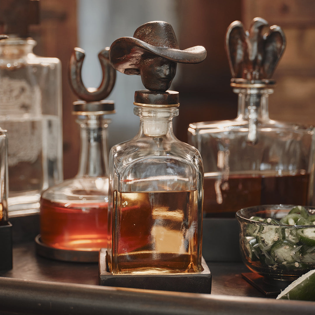 A cowboy iron decanter topper with vintage style glass bottle filled with liquor and iron coaster. At the background are more decanters with iron toppers.
