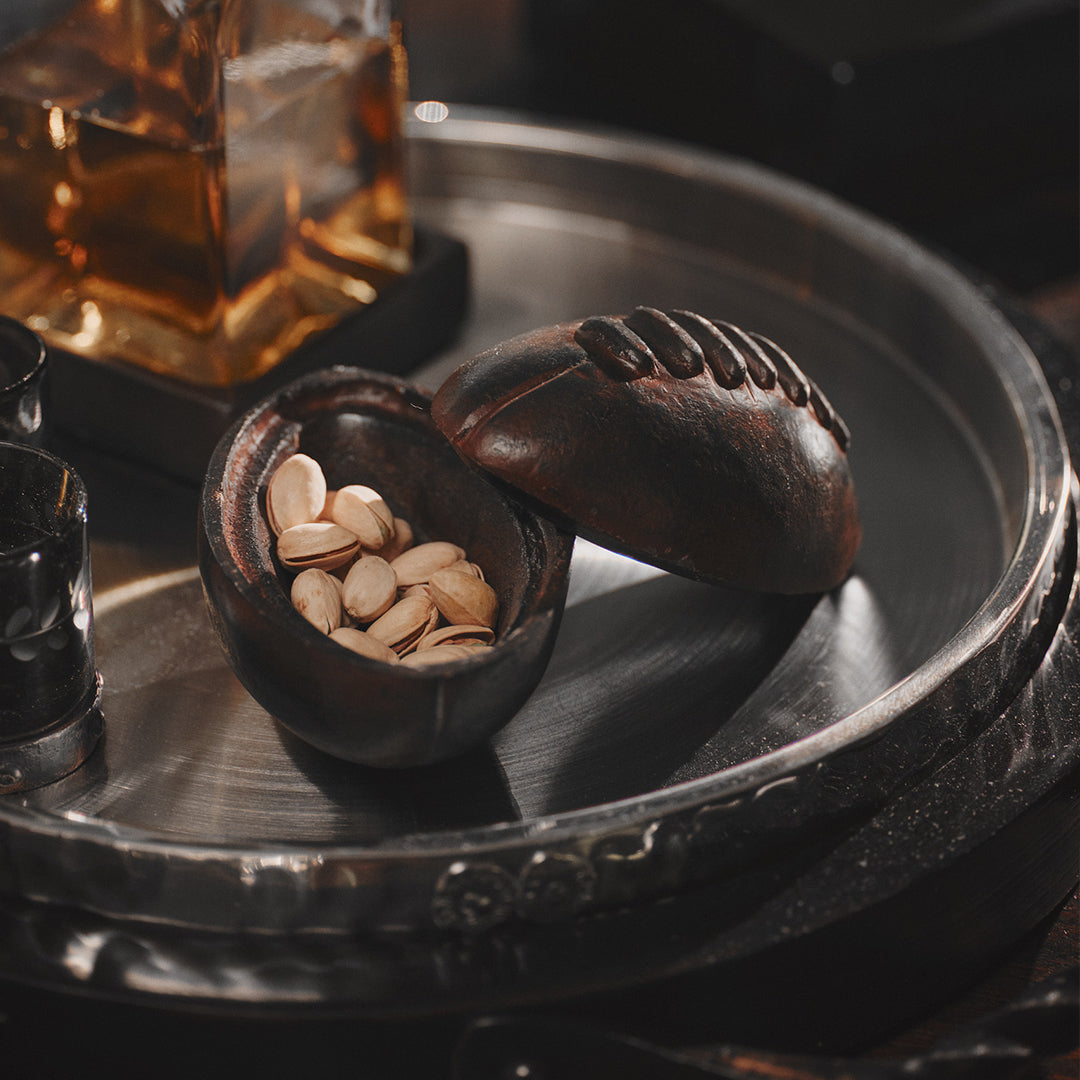 A tray with a iron football box with thee lid on its side, inside are pistachios. Behind it is a bottle.