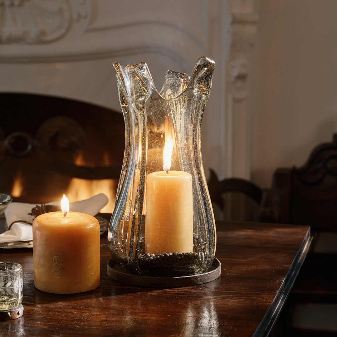 A wood table edge with a glass vase on a iron vase with a lit yellow candle. next to it is a lit yellow candle. On the background is a white chimney.