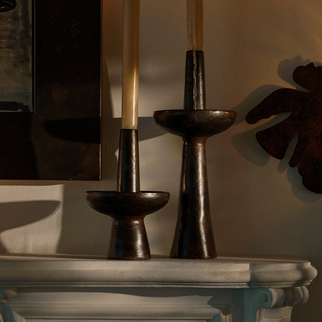 Two iron primitive and sculptural candlesticks with a candle each. On top of a white surface.