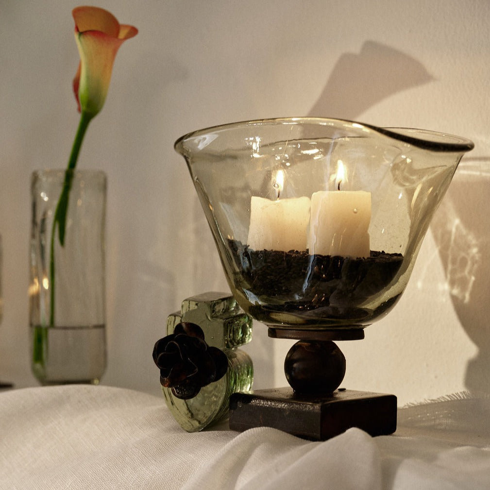 Two candles inside a fan-shaped vase resting atop a balin stand. On top of a white surface next to a flower inside a vase.