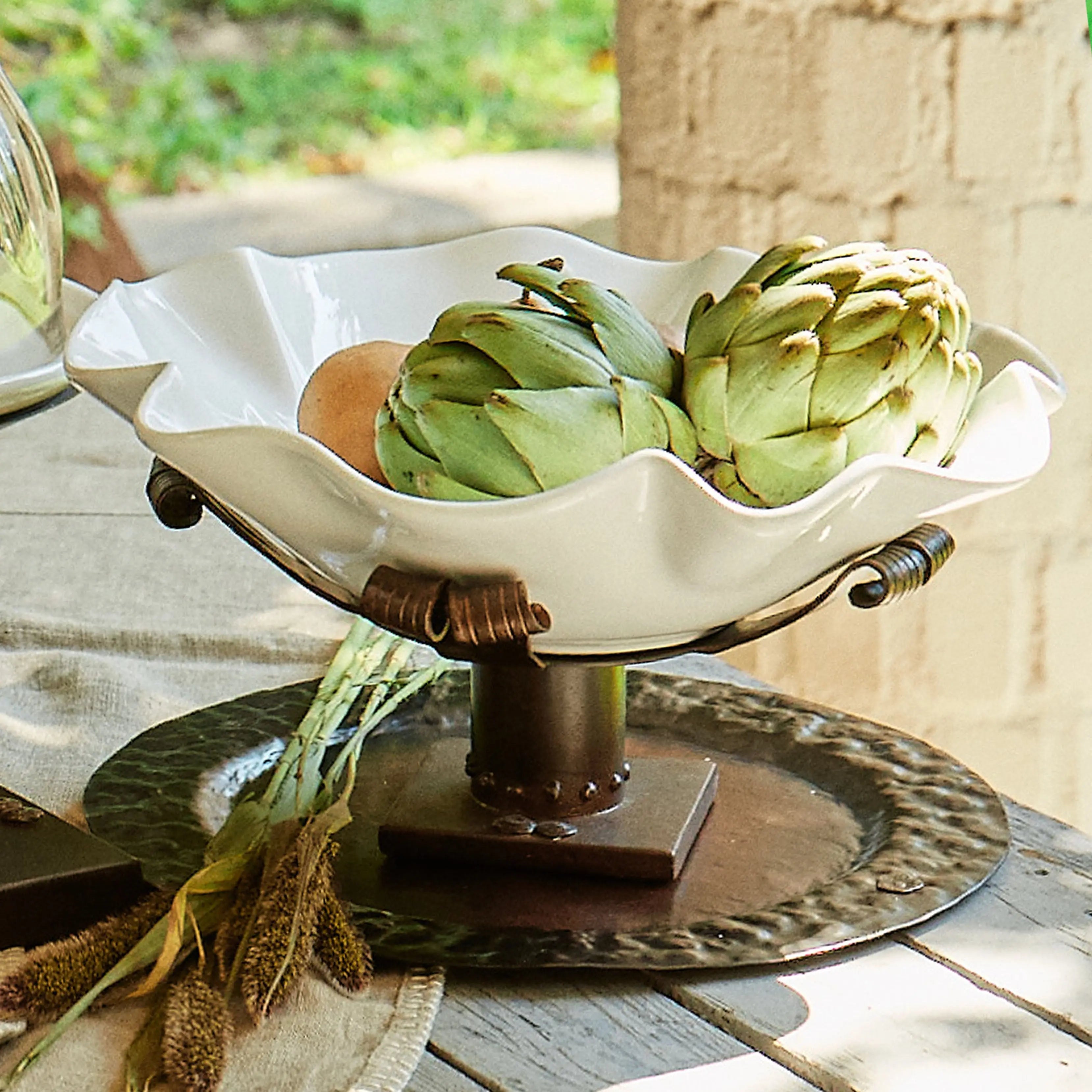 A white ceramic bowl with artichokes inside and a iron stand. On top of a iron tray in a light colored wood surface. Outside background.