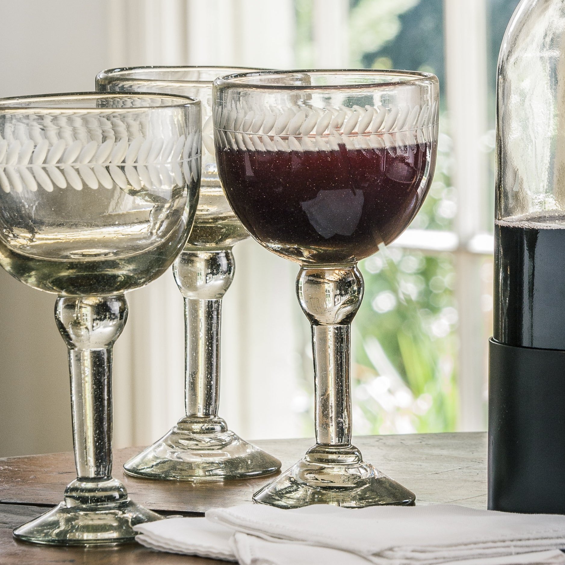 Three clear mouth blown goblets with laurel engravings. One is filled with a deep red liquid. Next to them is a bottle with dark liquid. On the background is a window.