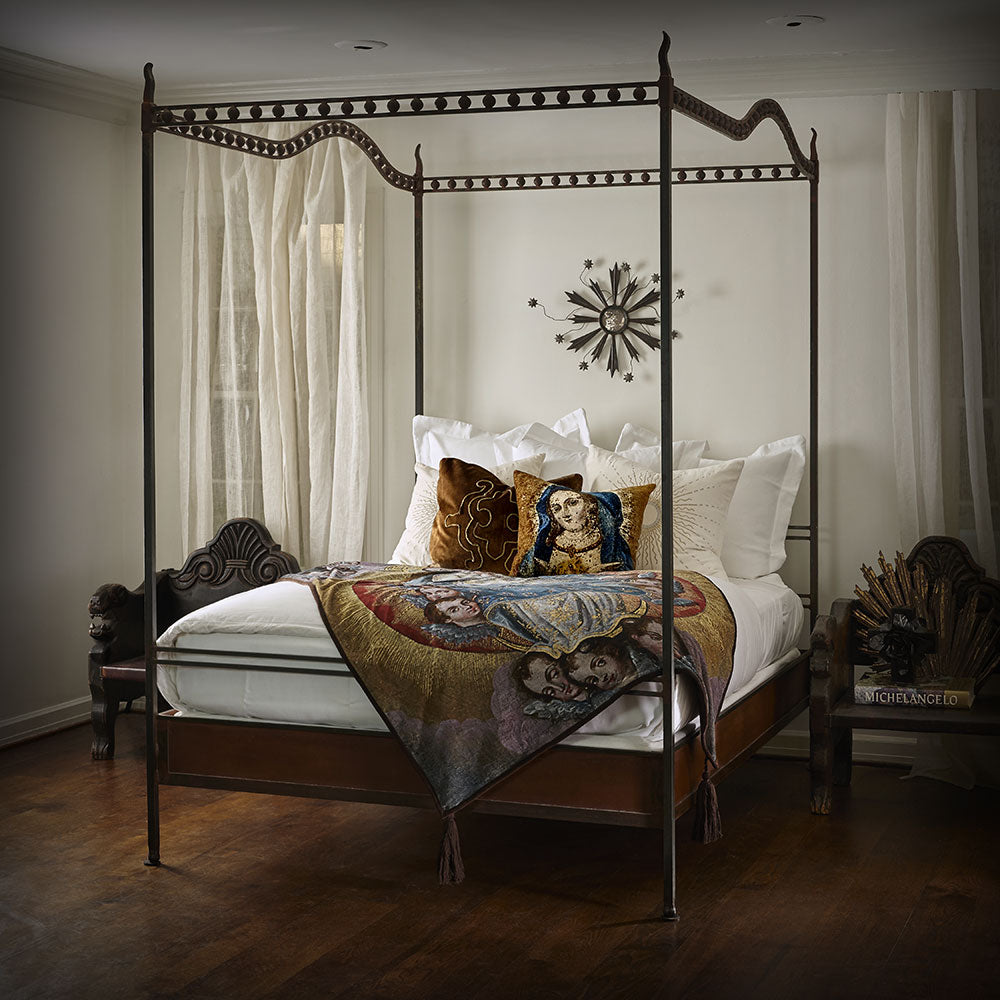 A canopy bed with white pillows and sheets- Inside a room with white walls and wood floor. On top of the bed is a mantel with faces and a pillow with the face of the virgin.