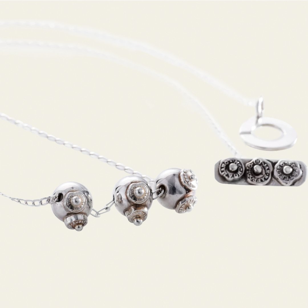 A delicate silver chain necklace with three ornate spherical charms featuring intricate designs. Alongside a rectangular charm engraved with seals featuring the brand logo.