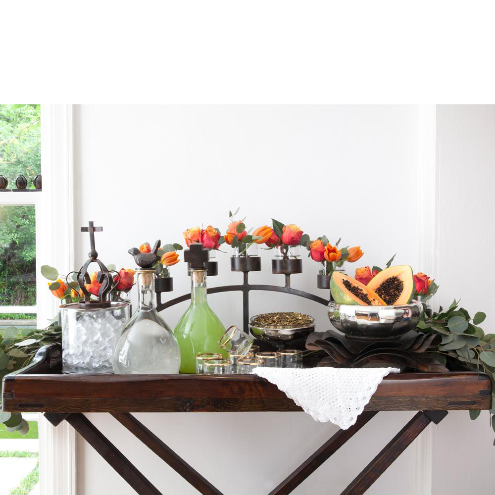 A candelabra with hand forged base and 9 glass vessels with orange flowers in each. Surrounded by glass bottles with ice and drinks, drinking glasses and owls with seeds and fruits. Everything is on top of a table against a white wall and a window.
