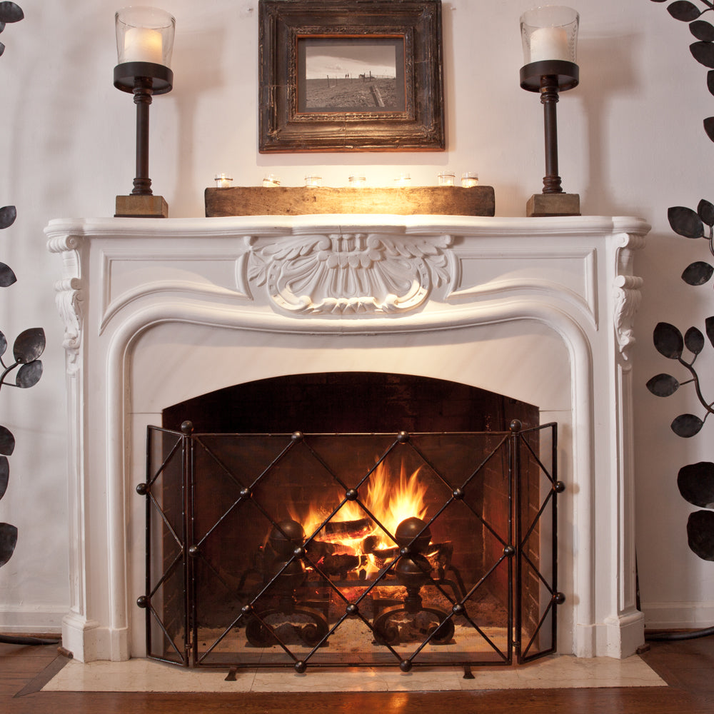 A white chimney with fire. A forged iron fire screen in front. Two iron stands with candle on top and a picture in the middle.
