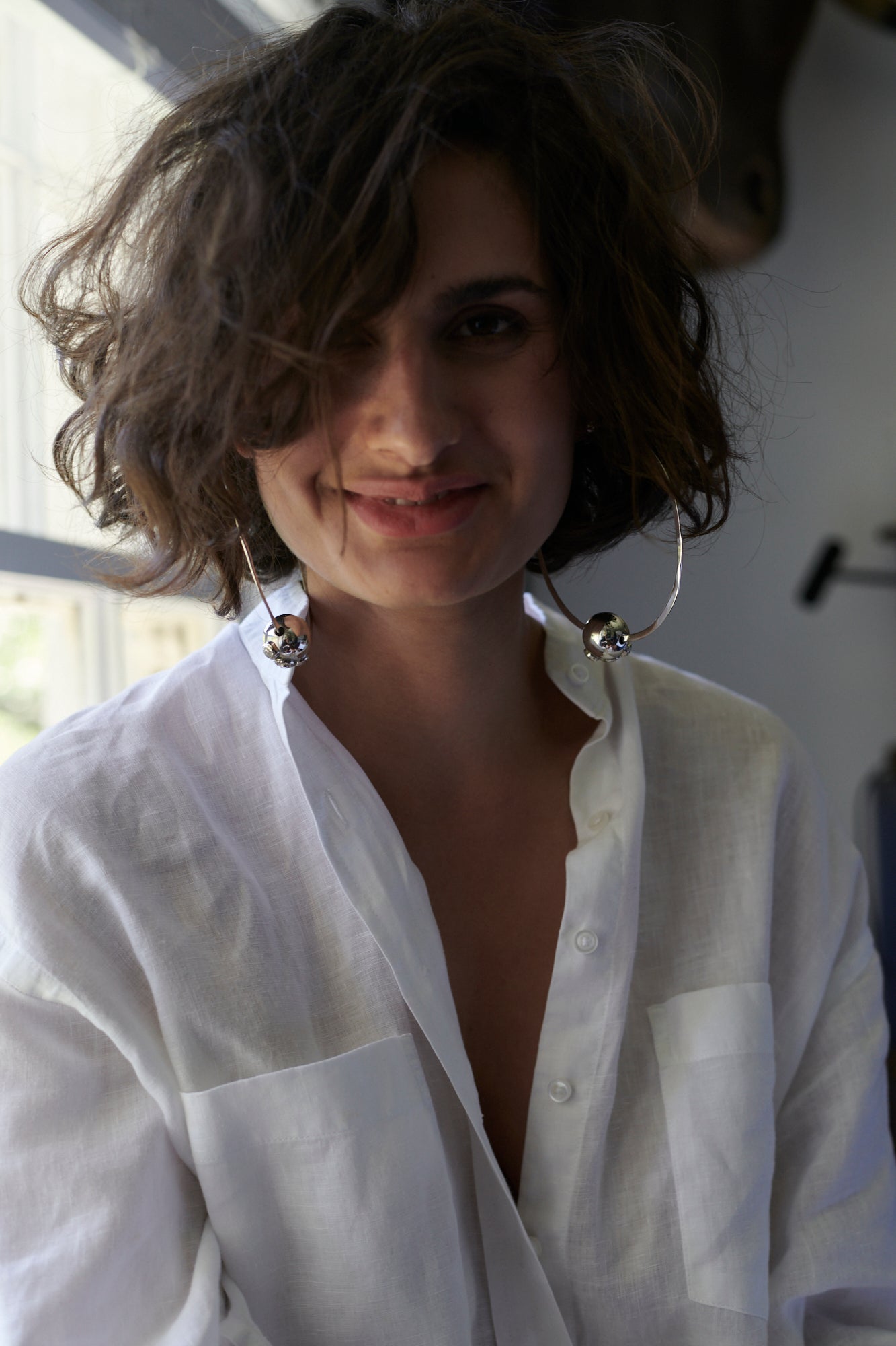 A brunette short haired woman wearing a white button up shirt and two big silver hoops with a silver bead in the center.
