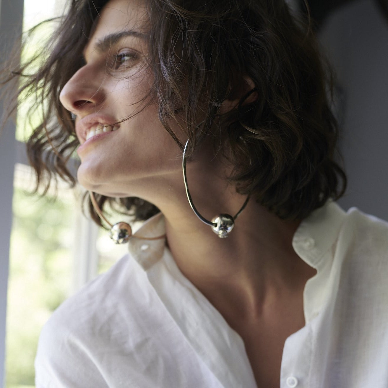 A short haired woman looking to the side wearing a white shirt and two big silver hoop earring with a silver bead.