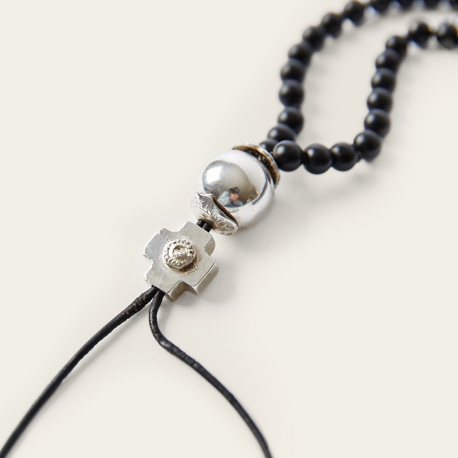 A necklace with black beads and at the end a silver ball and a silver cross followed by two black leather strings. Plain white background.