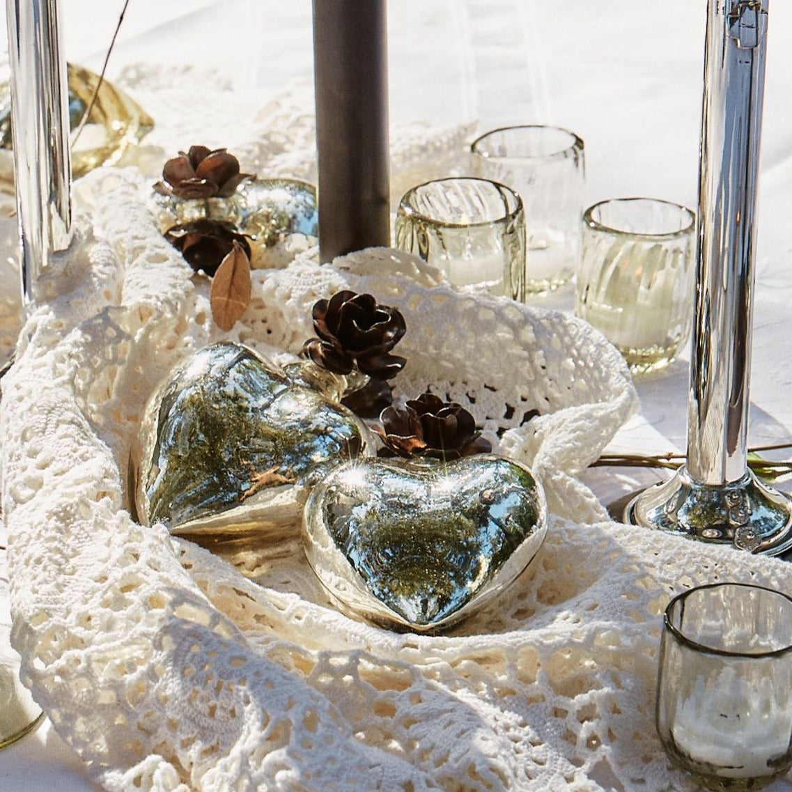 Three mouth blown puffed glass hearts with platinum finish and tiny iron rose on top of a white fabric. Surrounded by glasses.