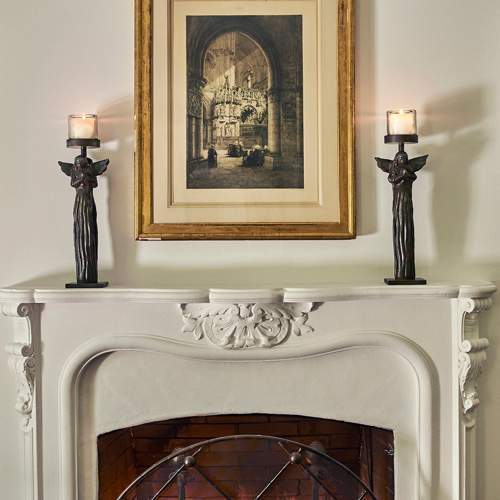 Two wax cast iron double angel on stand with mouth blown glass insert with lit candles on top of a white chimney. A photo in the center.