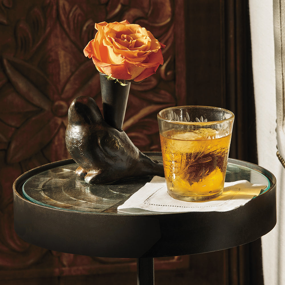 A mirror round table. On top of the table is a iron bird with a rose and glass with a yellow liquid. On the background is a carved wood.
