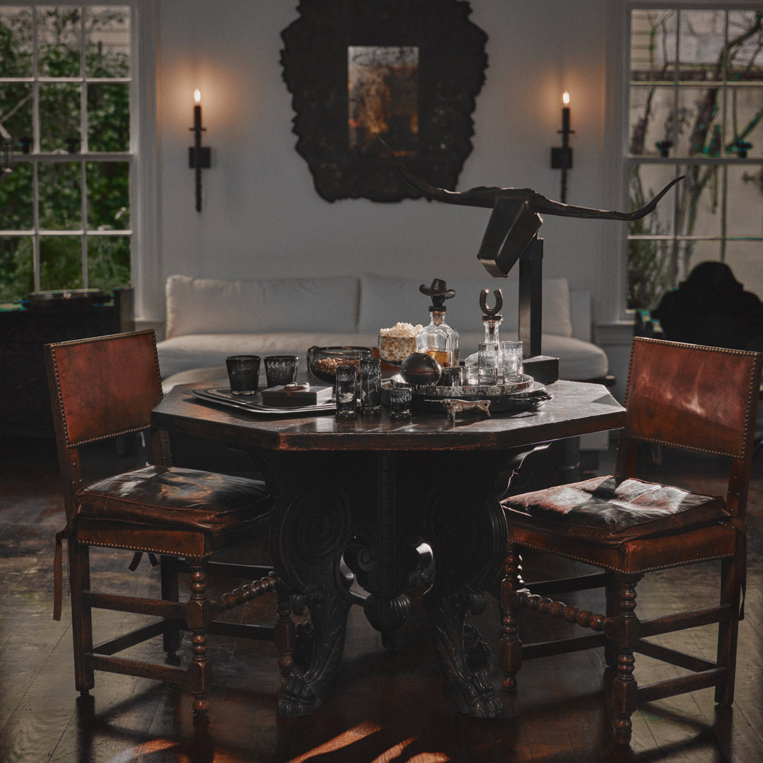 A wood table wit two brown chairs looking at each other from opposite sides. On too of the table are several glasses, an iron bull head decoration on a stand, some decanters and a tray. On the background is a white couch, two windows, two torches and a dark frame.