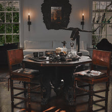 A wood table wit two brown chairs looking at each other from opposite sides. On too of the table are several glasses, an iron bull head decoration on a stand, some decanters and a tray. On the background is a white couch, two windows, two torches and a dark frame.