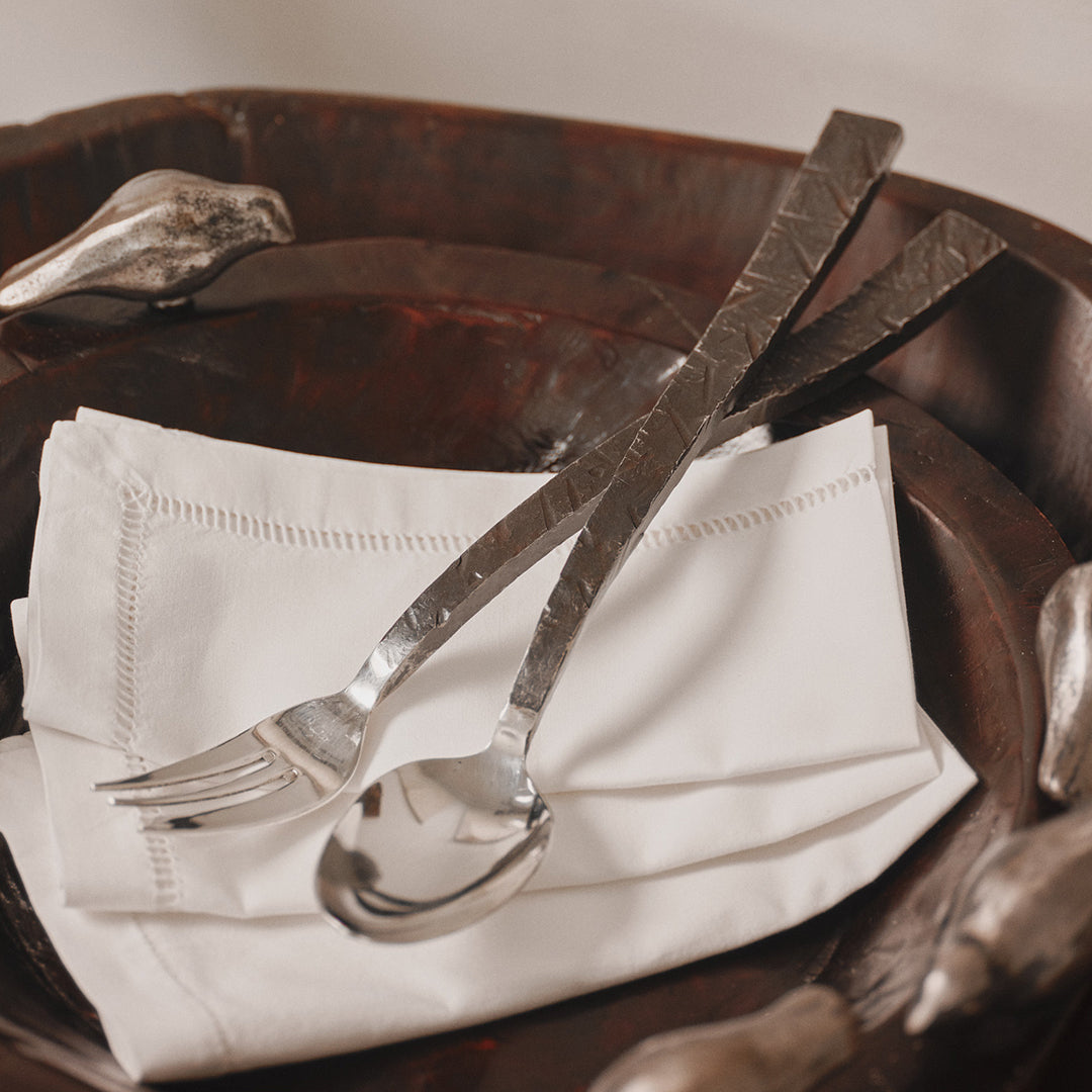 A pair of rustic cast iron handles with stainless steel serving spoon and fork on top of a white fabric napkin and a wood owl with a silver bird.