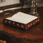 A wax cast iron square napkin holder with beaded detail trim. White napkins inside of it. It is resting on top of an iron surface.