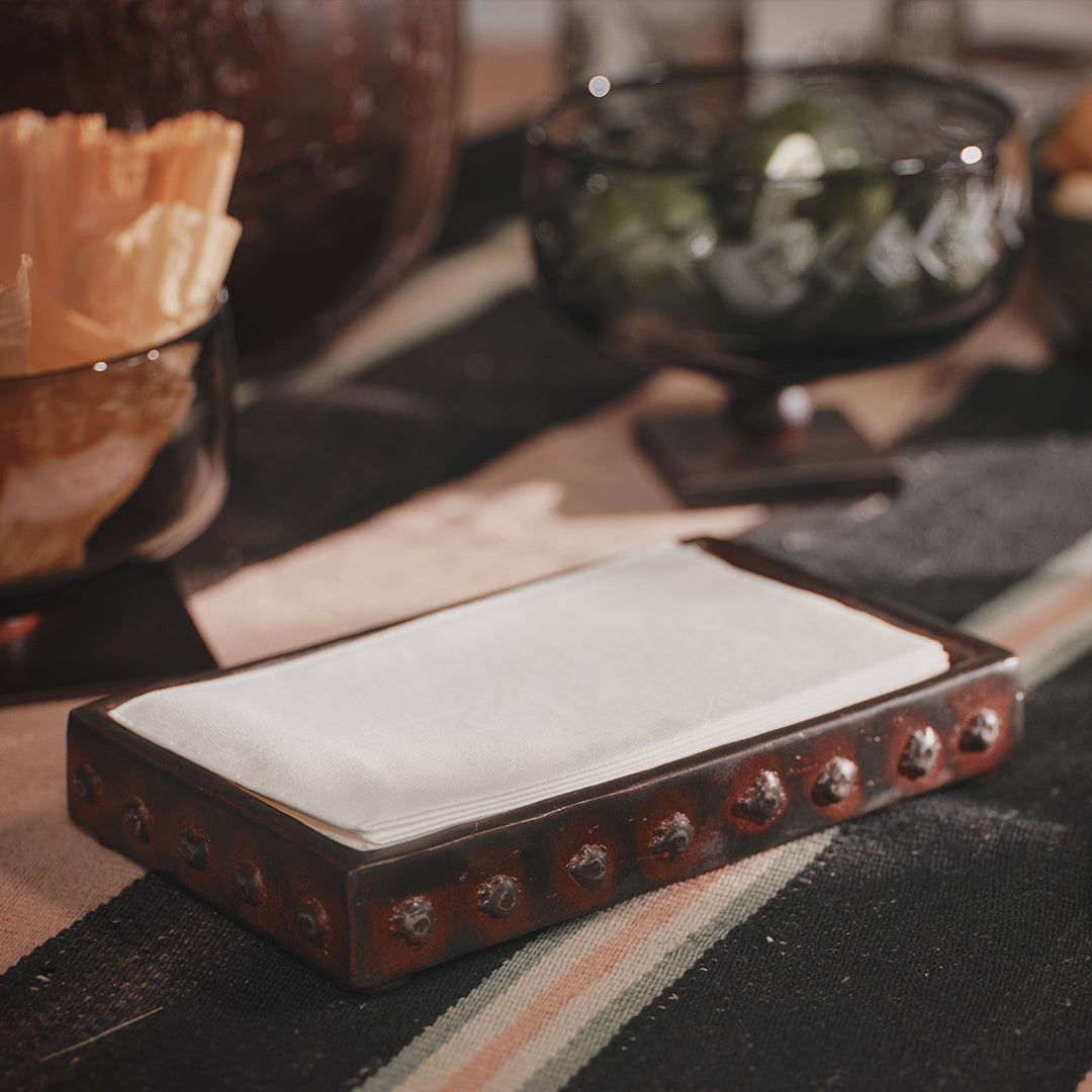 Wax cast iron rectangle napkin holder with beaded detail trim with white napkins inside it. Positioned on top of a mantel in front of two bowls.