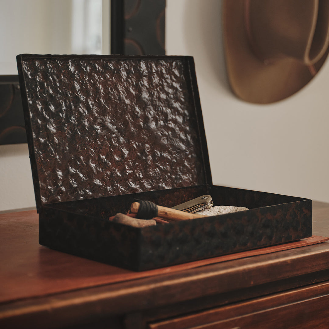 A hammered iron open box with some objects inside. The box is on top of a wood furniture and behind it a part of a hat can be seen.