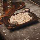 A hand carved small rectangular primitive wood bowl with two cast iron birds filled with pistachios on top of a grey surface.