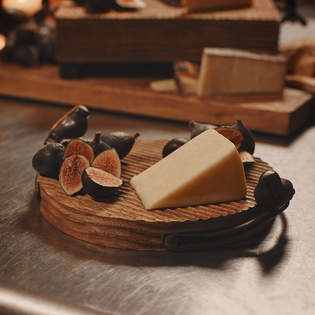 A rough-hewn round cutting board with cast iron bird handles. On the board are some figs and a cheese.