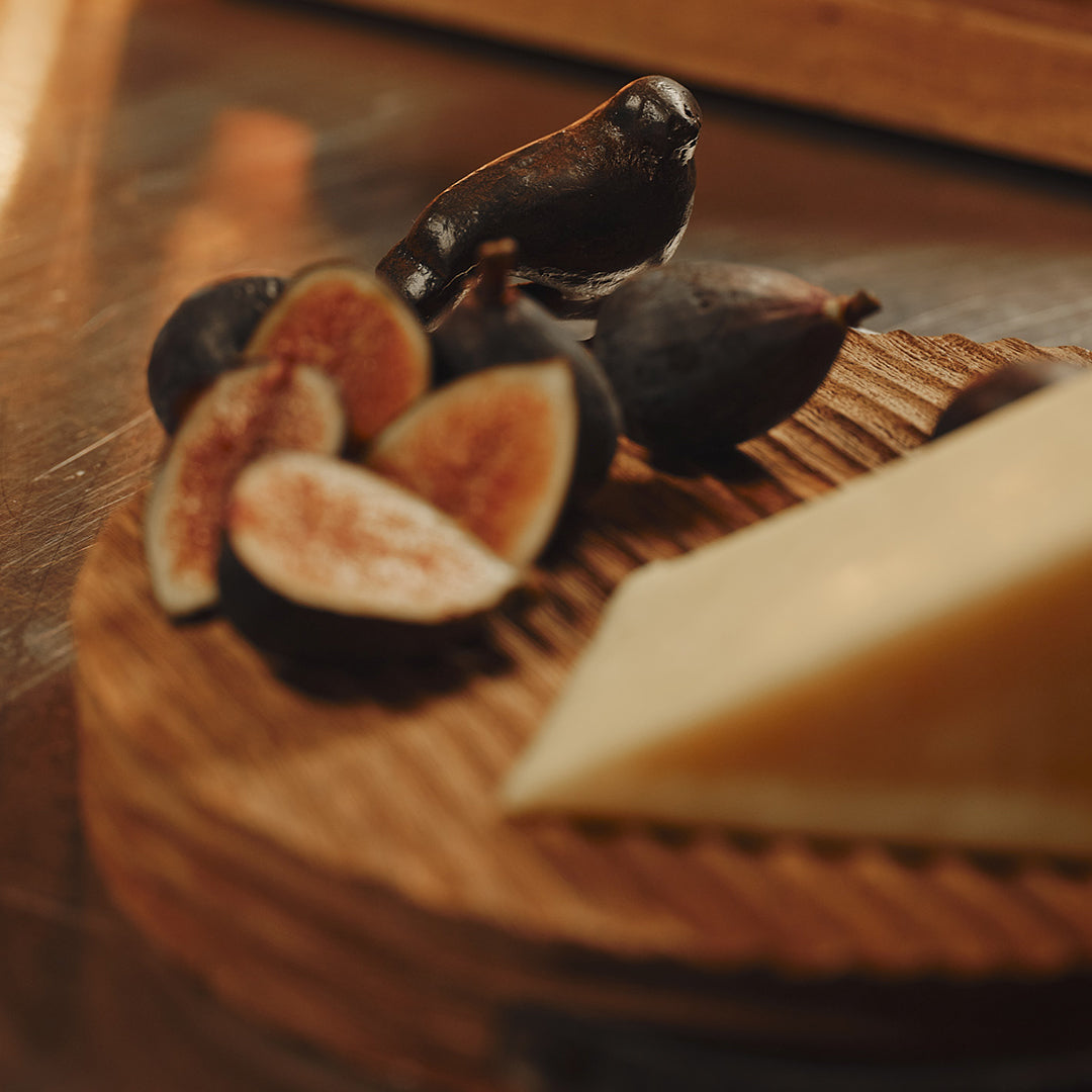 A  round cutting board with cast iron bird handles. On the board are some figs and a cheese.
