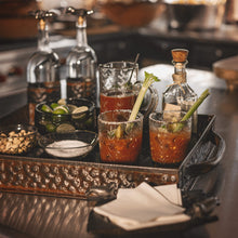 A hammered ottoman tray with  three glasses with cocktails and a celery, three bottles and bowls with lemons, salt and nuts.