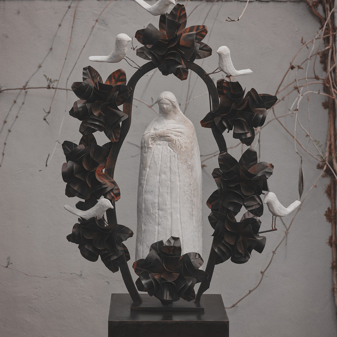 A hand carved stone statue representing the spirit of the virgin of  Guadalupe surrounded by an aura and halo of iron flowers and stone birds.  On the background is a light grey wall and brown vines.