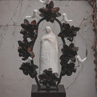 A hand carved stone statue representing the spirit of the virgin of  Guadalupe surrounded by an aura and halo of iron flowers and stone birds.  On the background is a light grey wall and brown vines.