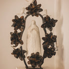 A hand carved stone statue representing the spirit of the virgin of  Guadalupe surrounded by an aura and halo of iron flowers and stone birds.  White wall background.