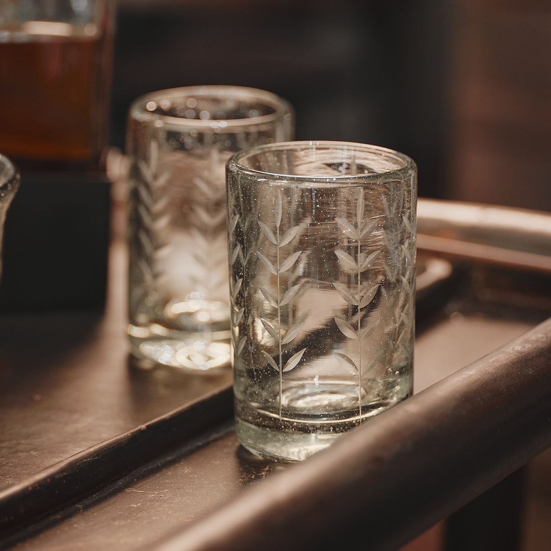 Two small mouth blown glasses with vertical laurel branches. On top of a dark brown table.