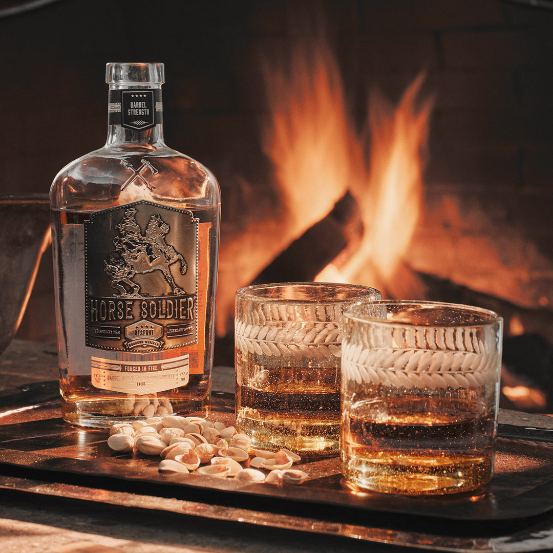 A tray with two glasses and a alcohol bottle and some pistachios. On the background is a fire.