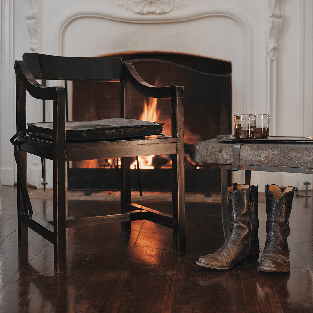 On the background is a white  lit chimney. A wood chair next to a table that holds a tray and two glasses with a liquid. Under the table is a pair of boots.