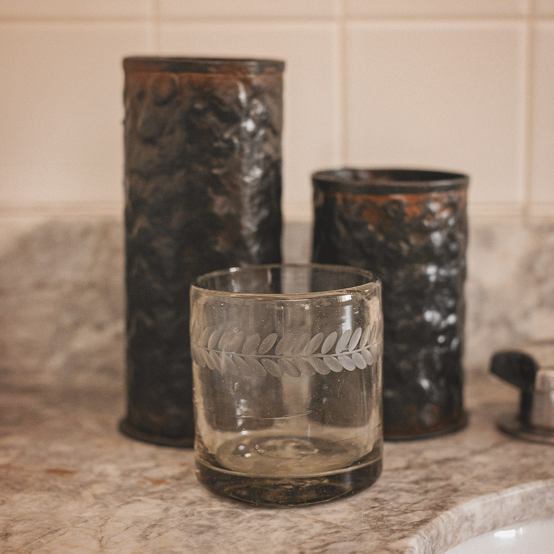 A clear glass with a laurel engraving. Behind are two hammered iron vases.