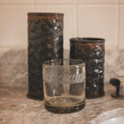 A clear glass with a laurel engraving. Behind are two hammered iron vases.