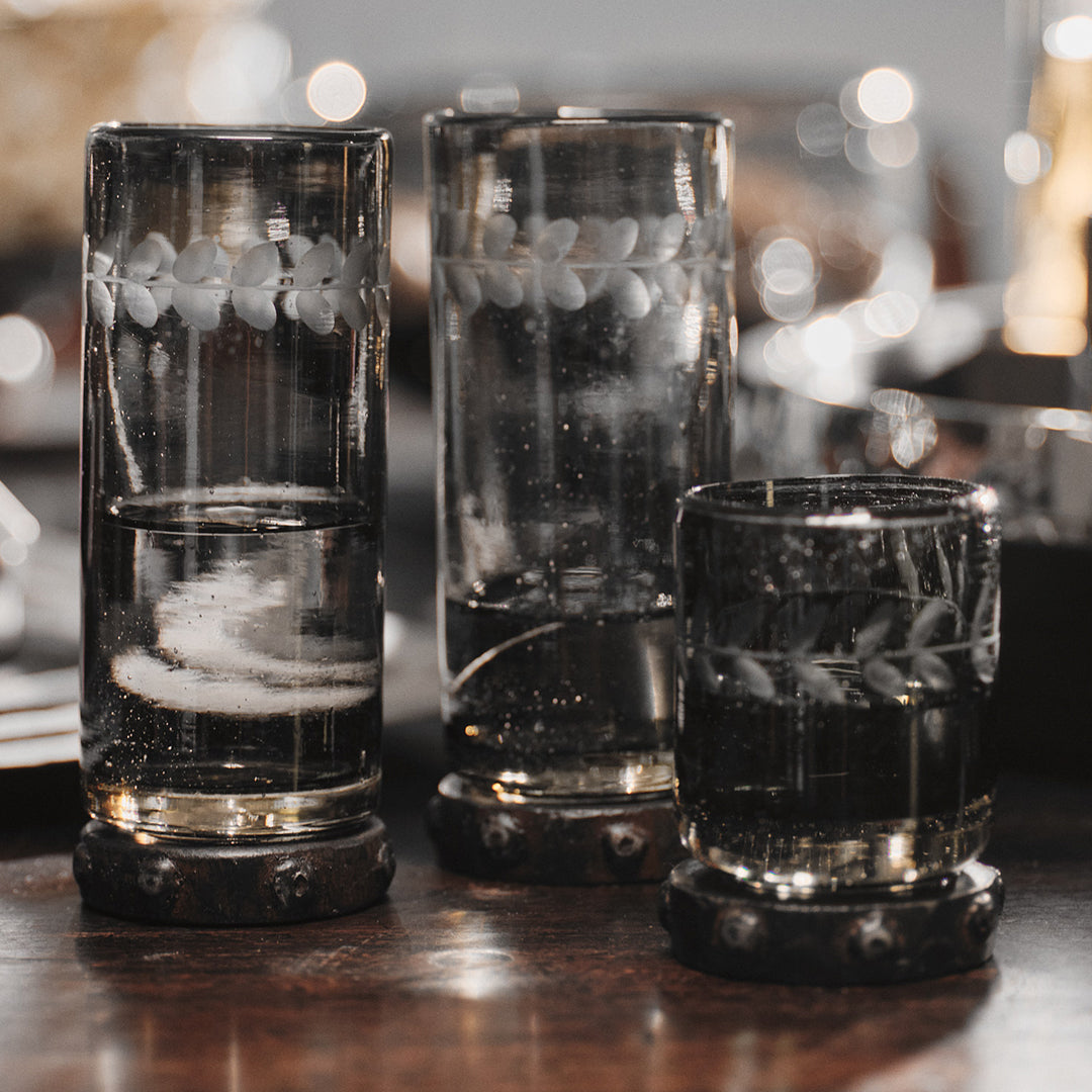 A mouth blown and engraved smoke colored shot glass with beaded iron base next to two  mouth blown and engraved smoke colored tall glasses with beaded iron base.
