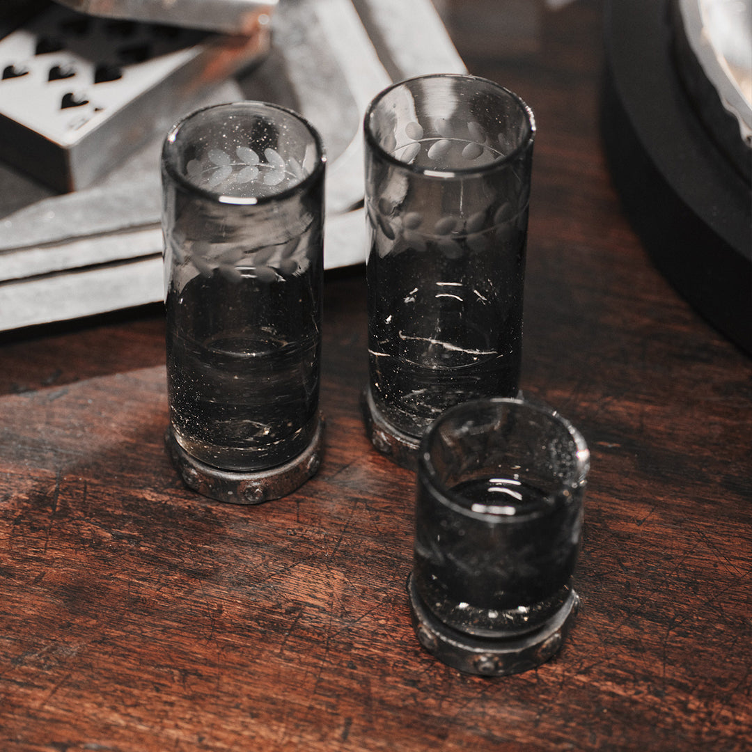 A mouth blown and engraved smoke colored shot glass with beaded iron base next to two  mouth blown and engraved smoke colored tall glasses with beaded iron base. Photo taken from a top view. On the background are some playing cards.