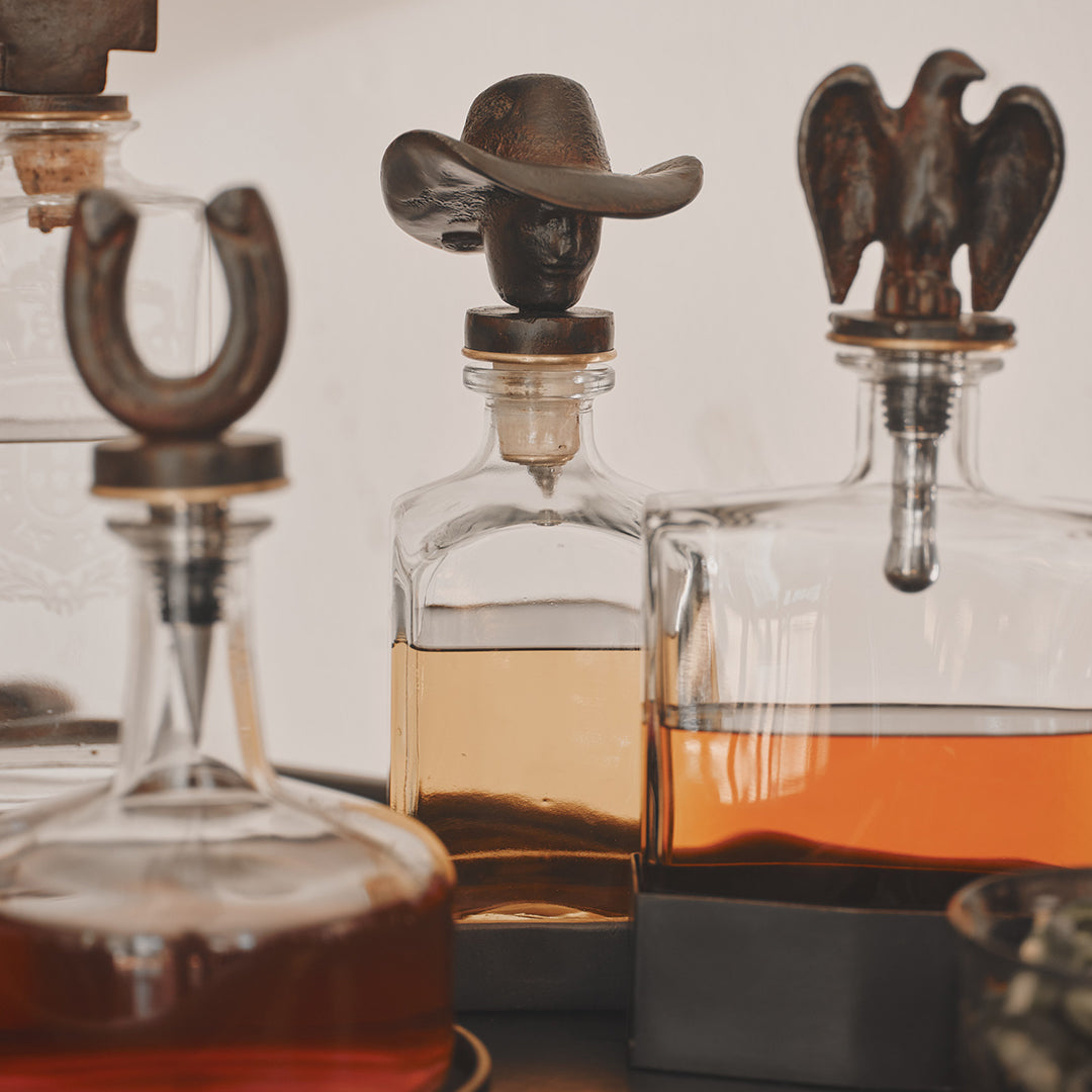 Three decanters with iron toppers. One is a cowboy, another a eagle and a horseshoe. White background.