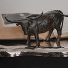 A longhorn bull iron sculpture on top of a silver surface and a opened book behind it.