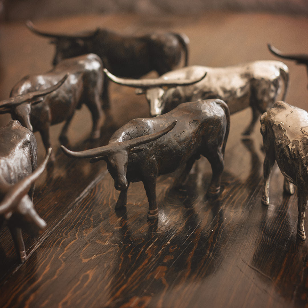 Several  longhorn bull iron sculptures and other longhorn bull nickel plated sculptures on top of a wooden table.