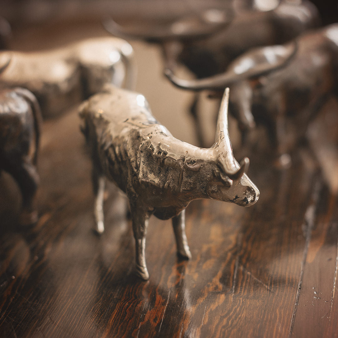 A  nickel plated longhorn bull  iron sculpture in front of other iron longhorn sculptures. Wood floor.
