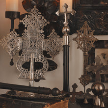 A nickel plated cross with natural iron handle supported on a gallery stand. in the middle of other objects like a silver cross and others. In front is another cross on a stand laying down.