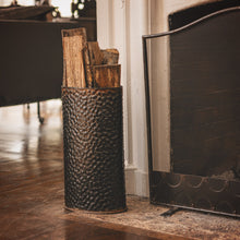 A standard oval hammered iron  vessel filled with logs next to a chimney. Wood floor.