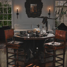 A dark table with two brown chairs facing each other. On the table are some trays, glasses, bowls, decanters and a iron bull's head sculpture. On the background is a white wall with two windows, two lit torches, a white sofa and a dark frame.