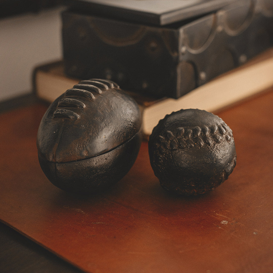 A iron football ball and a iron baseball  ball next to each other on top of a wood surface.